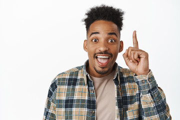Portrait of happy african american guy saying great news, raising finger up and smiling, laughing excited, having awesome idea, suggesting plan, white background