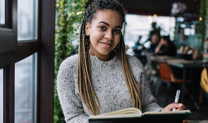 Portrait of beautiful female student with braiding Afro hairstyle sitting at cafeteria with education textbook for learning information and looking at camera, clever hipster girl studying indoors
