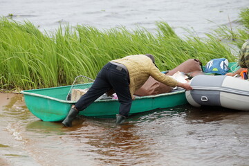 A man without lifejacket in warm clothes and rubber boots pushes a loaded rowing and inflatable...