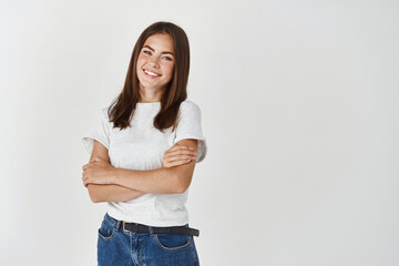 Beautiful young woman looking happy and carefree, standing with arms crossed on chest over white...