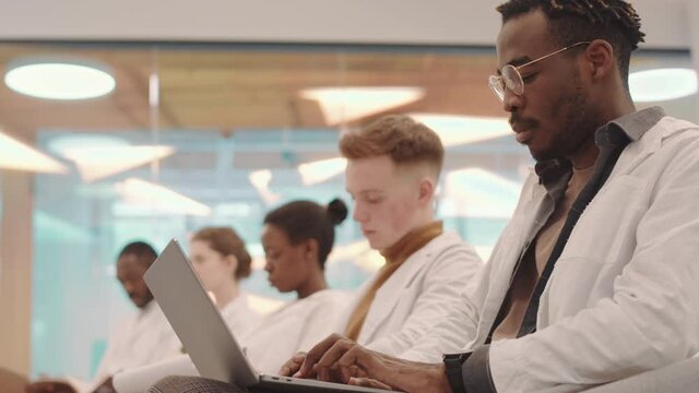 Side-view medium slowmo of young Afro American medical student and his multiethnic classmates sitting in row in modern auditorium taking notes on laptop during lecture
