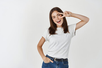 Happy brunette woman showing peace or victory sign on eye and smiling, express joy and excitement, standing in t-shirt against white background