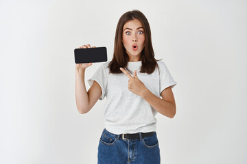 Impressed brunette woman showing mobile phone screen, pointing at cellphone and looking amazed at camera, white background