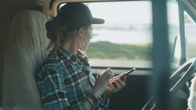 Young Caucasian woman in casualwear using smartphone while sitting in van on driver seat