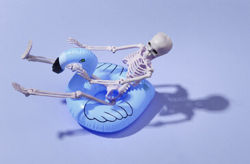 Toy skeleton with inflatable flamingo on purple bright background. Halloween theme. Beach vacation concept. Summer rest.
