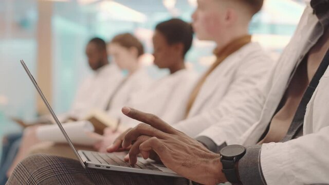 Close-up of unrecognizable Black male hands of medical student typing lecture notes on laptop with group of other students sitting in row in blurred background
