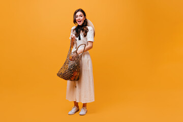 Young girl in midi dress with straw hat on her back is putting orange into string bag on isolated background