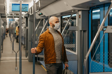 Fototapeta na wymiar A man with a beard in a medical face mask to avoid the spread of coronavirus is riding a modern subway car. A bald guy in a surgical mask is keeping social distance on a train.