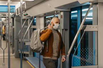 A man with a beard in a face mask to avoid the spread of coronavirus is putting on a gray backpack while riding a subway car. A bald guy in a surgical mask is keeping social distance on a train.