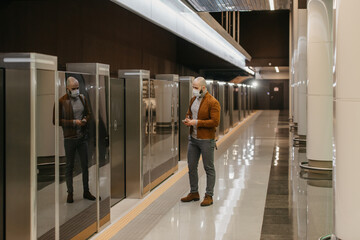 A man in a medical face mask to avoid the spread of coronavirus is scrolling the news on a smartphone while waiting for a train on the subway. A bald guy in a surgical mask is keeping social distance.