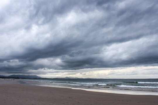 Photograph that looks like a painting - perspective of Byron Bay Australia beach with mountains around the bay and dark storm clouds rolling overhead.