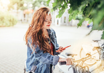 Portrait of smiling at camera red curled hair caucasian teen girl unlocking bike at Bicycle sharing...