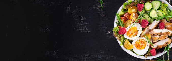 Grilled chicken meat and fresh vegetable salad of tomato, cucumber, egg, lettuce and raspberry. Ketogenic diet. Buddha bowl dish on dark background. Top view, banner