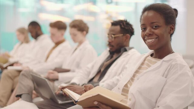 Side-view medium slowmo portrait of smiling female Afro American medical student taking notes in copybook then posing for camera sitting in row with classmates in modern auditorium having lecture