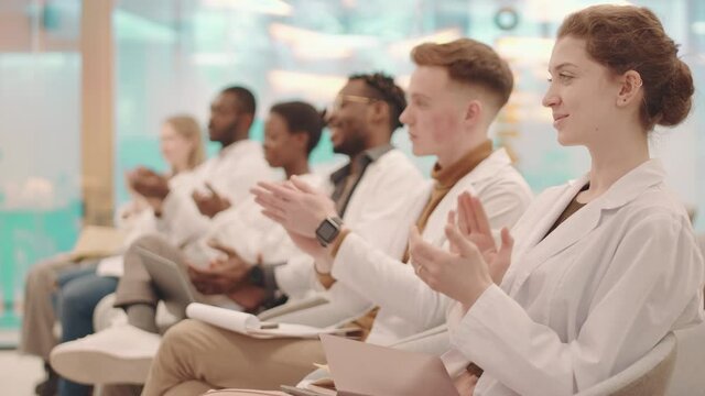 Side-view medium shot of group of young multiethnic medical students in white coats applauding to professor sitting in row in modern lecture hall