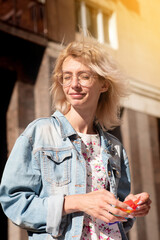 a beautiful woman in glasses and a denim jacket stands against the background of a cityscape with a pop-it in her hands with her eyes closed looking towards the sun. Vertical photo