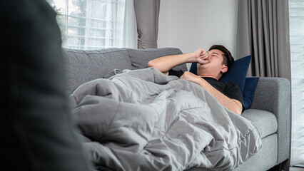 Asian man is lying with blanket on couch at home to rest when having high fever, headache and using