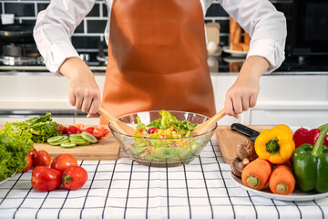 Asian housewife wearing apron and using ladle to mixing vegetable salad in bowl while standing preparing
