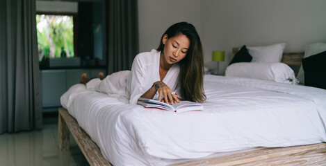 Calm Asian female lying on bed and reading book