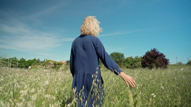 A European woman, middle-aged in a dress, walks on a prak with tall grass. With a philosophical romantic mood. Slow motion