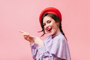 Brunette girl with red lipstick is smiling and pointing her fingers to left. Portrait of woman in beret with place for text on pink background