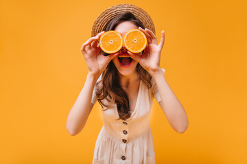 Girl in white dress and hat covers her eyes with oranges and poses on isolated background