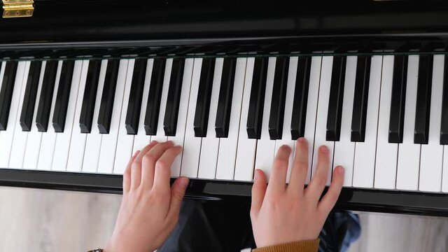 Close up of the hands of a young child playing the piano. Close up top view of a youngster practicing a song on the keys of an upright shiny black piano, with subtle zoom in.