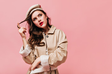 Woman in trench coat and beret looks thoughtfully up, bites her lip and winds her hair onto her finger