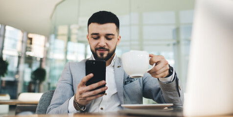 Fototapeta na wymiar Positive businessman with smartphone and cup of coffee in office