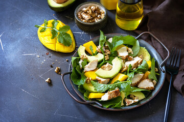Healthy diet salad with arugula, mango, avocado, chicken grill, walnuts and vinaigrette dressing on a dark stone countertop. Copy space.