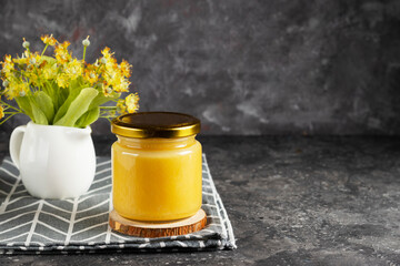 Summer linden honey in an open glass jar with a wooden honey spoon with a bouquet of linden flowers in a white milk jug on a gray napkin copy space