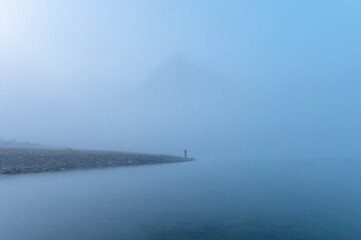 Traveler man standing alone in blue fog with rocky mountains by the lake in the morning