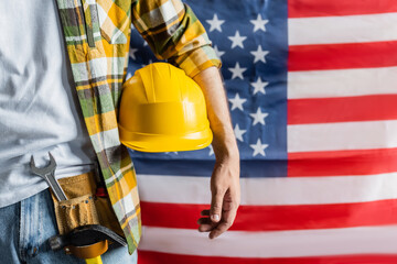 cropped view of workman in plaid shirt and tool belt holding hardhat near blurred usa flag, labor day concept - obrazy, fototapety, plakaty