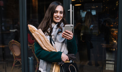 Fototapeta na wymiar Delighted trendy woman near entrance of pastry shop with smartphone and bread