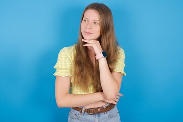 Fototapeta premium Dreamy young beautiful blonde woman standing against blue background with pleasant expression, looks sideways, keeps hand under chin, thinks about something pleasant.