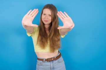 Portrait of smiling young beautiful blonde woman standing against blue background looking at camera and gesturing finger frame. Creativity and photography concept.