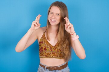 young beautiful blonde woman standing against blue background holding fingers crossed with worried expression hoping boss didn't noticed mistakes at work.