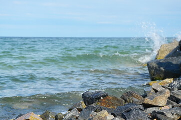 Close-up, waves break on the stone coast of the sea.
On the horizon are blue skies and small clouds.