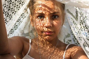 Mysterious portrait of a beautiful mestizo young girl with light shadow pattern on face. Adorable charming green eyed female looking at camera. Creative art shadows from white lace sheet at sunny day.
