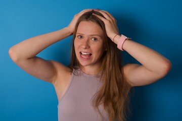 Cheerful overjoyed young beautiful blonde woman standing against blue background reacts rising hands over head after receiving great news.