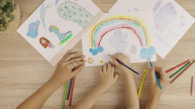 Top view seeing hands and arms of mother and her daughter kid girl are drawing and coloring beautiful rainbow pictures on white paper shows concept of parental time for happiness together activity.