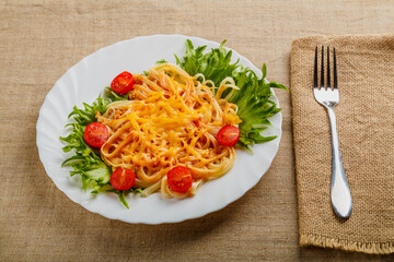 A plate of pasta with cheese and tomatoes decorated with herbs on a wooden table next to a linen napkin and a fork.