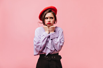 Nice girl dressed in purple blouse, red beret and pants posing offendedly on pink background