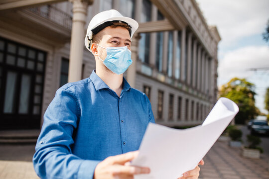 Architect looking at blueprints in a building site. Portrait of an architect builder in protective mask and helmet studying layout plan of the object.