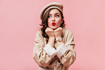 Green-eyed woman in trench coat and hat folded her lips into tube and posing on isolated background