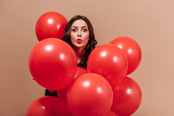Fototapeta na wymiar Green-eyed girl looks at camera in surprise, holding red balloons on beige background