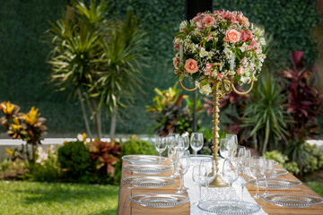 Arreglo floral sobre una mesa de madera con cristalería montada en un jardín para un evento social en Mexico