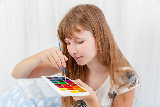 Blonde girl picks up with a brush the color of paint on a watercolor palette. homework. education. Creativity, drawing