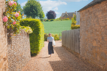 Fototapeta na wymiar A young blonde female tourist explores a quaint country lane in the rural English countryside village of Chipping Campde, on a summer day in the Cotswolds, Gloucestershire, UK.