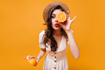 Beautiful green-eyed dark-haired girl in hat and sundress sends kiss and poses with orange halves